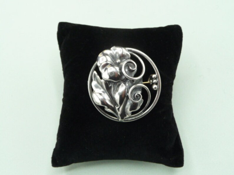 May include: A silver-toned floral brooch with a flower and leaf design, set within a circular frame. The brooch is displayed on a black velvet pillow against a light green background. The brooch has a pin closure on the back.