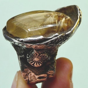 May include: A handcrafted ring featuring a large, oval-shaped, golden-brown gemstone with internal inclusions, set in a textured silver and bronze metal band. The ring has a unique, organic design with a floral accent.