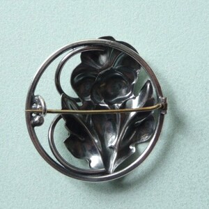 May include: A silver-toned brooch featuring a floral design within a circular frame. The brooch showcases a detailed flower and leaves, with a pin closure on the back. The design is intricate and the metal has a polished finish.