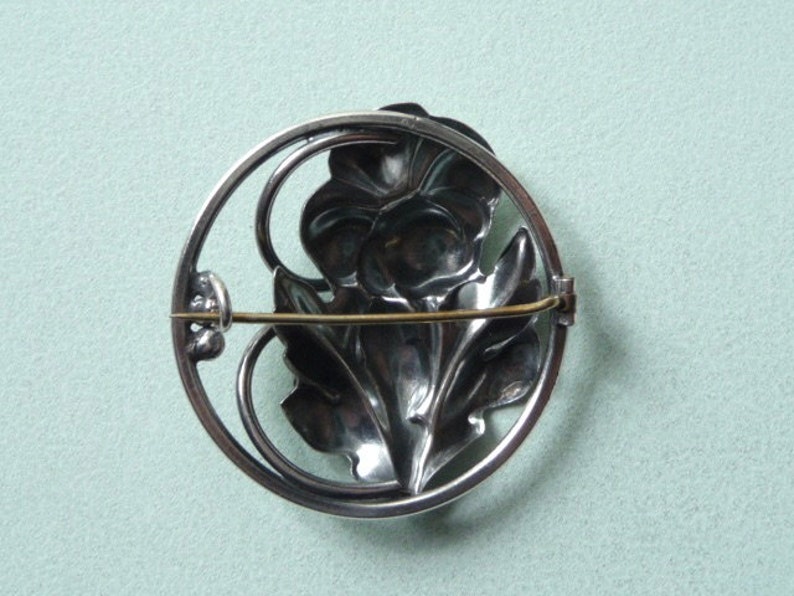 May include: A silver-toned brooch featuring a floral design within a circular frame. The brooch showcases a detailed flower and leaves, with a pin closure on the back. The design is intricate and the metal has a polished finish.
