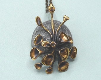 May include: A bronze-toned pendant necklace featuring a stylized floral design. The pendant has a cluster of petal-shaped elements in a warm gold color, set against a dark gray, textured stone. The chain is a matching bronze color.