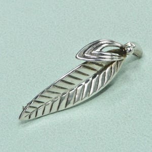 Peut inclure: Broche argentée en forme de feuille, avec des nervures détaillées et une boucle stylisée. La broche a une finition polie et est posée sur un fond bleu-vert clair. Ce bijou est un accessoire inspiré de la nature.