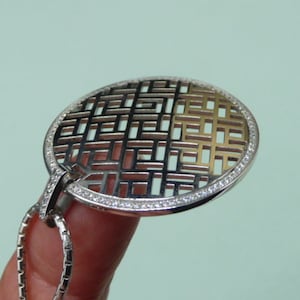 Peut inclure: Un collier avec un pendentif rond en argent et or, orné d'un motif géométrique. Le pendentif est entouré d'une rangée de petites pierres claires.