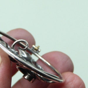 May include: A silver-toned brooch with a circular design. The brooch features a detailed floral or leaf pattern and a pin closure on the back. The metal has a polished finish, and the design is slightly raised, creating a three-dimensional effect.