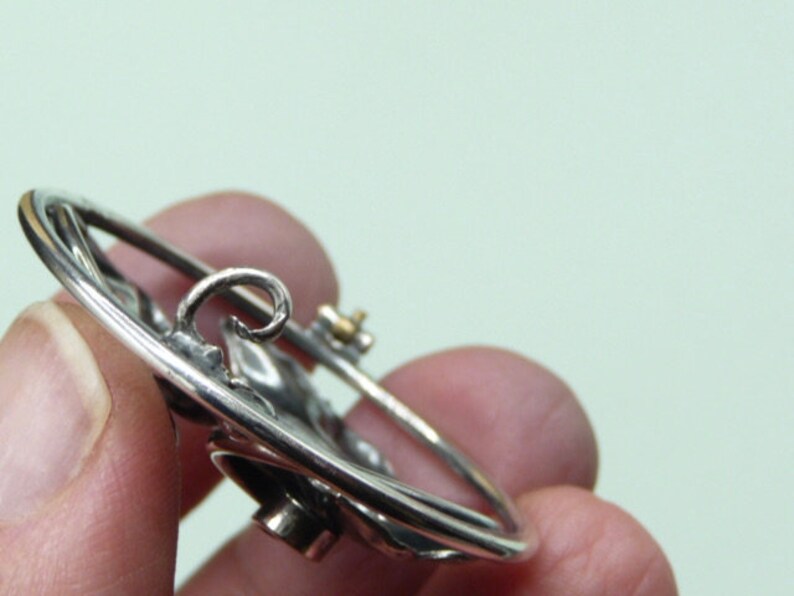 May include: A silver-toned brooch with a circular design. The brooch features a detailed floral or leaf pattern and a pin closure on the back. The metal has a polished finish, and the design is slightly raised, creating a three-dimensional effect.