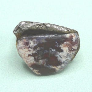 May include: A natural stone ring with a silver-colored metal band. The stone features a mix of burgundy, white, and dark gray patterns. The ring's design is organic and unique, showcasing the natural beauty of the stone.