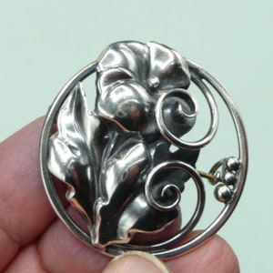 May include: A silver-toned brooch featuring a floral design within a circular frame. The brooch showcases detailed leaves, a central flower, and decorative swirls. The design includes small, round accents, creating an elegant accessory.