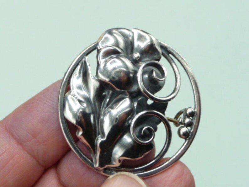May include: A silver-toned brooch featuring a floral design within a circular frame. The brooch showcases detailed leaves, a central flower, and decorative swirls. The design includes small, round accents, creating an elegant accessory.
