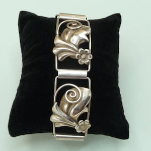 Peut inclure: Un bracelet en argent avec un motif floral. Le bracelet est composé de plusieurs maillons, chacun avec un motif floral différent. Le bracelet est posé sur un coussin en velours noir.