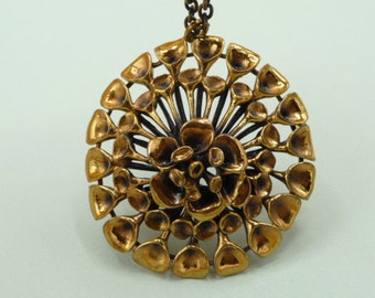 May include: A gold-toned pendant necklace with a circular design featuring a stylized flower with multiple petals. The pendant is made of metal and has a textured surface.