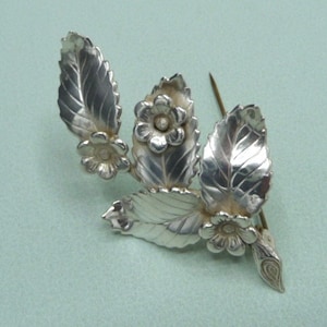 Peut inclure: Broche en argent avec un motif floral composé de trois feuilles et trois fleurs. Les feuilles sont texturées et les fleurs sont simples et rondes.