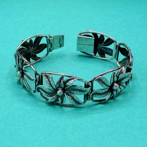 Peut inclure: Un bracelet à maillons en argent présentant un motif floral répétitif. Chaque maillon se compose d'un design de fleur à cinq pétales avec des textures de feuilles détaillées et un accent sphérique central. Fermoir à boîte rectangulaire.