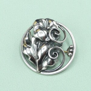May include: A silver-toned brooch featuring a floral design within a circular frame. The brooch showcases detailed leaves, a flower, and swirling accents. A small cluster of three beads and a gold-colored pin are also visible.