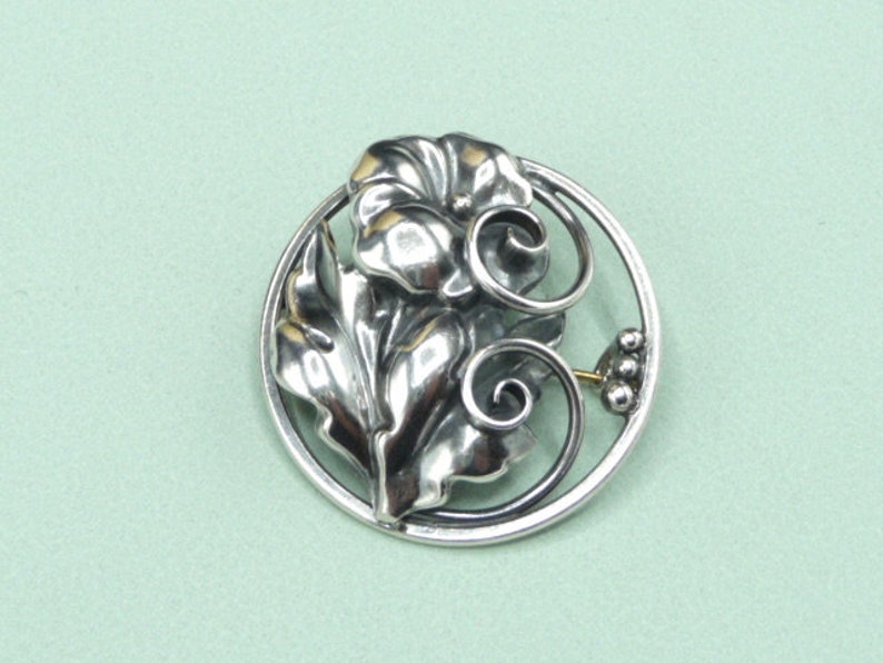 May include: A silver-toned brooch featuring a floral design within a circular frame. The brooch showcases detailed leaves, a flower, and swirling accents. A small cluster of three beads and a gold-colored pin are also visible.