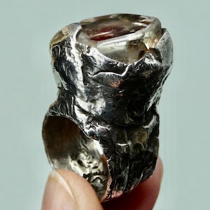 May include: A silver ring with a large, faceted, clear quartz stone with red inclusions. The ring band is textured and has a rough, organic look.