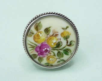 May include: A silver ring with a white porcelain top featuring a hand-painted floral design of yellow and pink roses with green leaves.