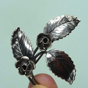 Peut inclure: Une broche en argent en forme de brin de fleur avec deux roses et trois feuilles.