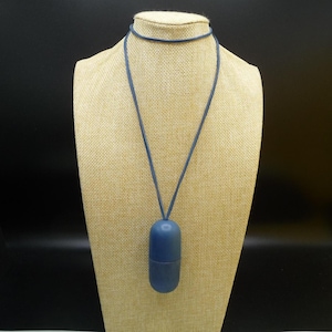 Könnte beinhalten: Eine blaue Anhängerkette mit einem zylindrischen Anhänger. Der Anhänger hat eine einfarbige blaue Farbe und scheint aus einem glatten Material zu bestehen. Die Halskette hat eine passende blaue Kordel. Die Halskette wird auf einer beigefarbenen Schaufensterpuppe präsentiert.