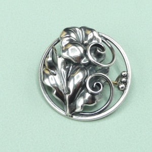 May include: A silver-toned brooch featuring a floral design within a circular frame. The brooch showcases detailed leaves and a flower with curled petals. A small cluster of beads adds a delicate touch to the design. The brooch is set against a light green background.