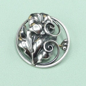 May include: A silver-toned floral brooch with a detailed design. The brooch features a central flower with layered petals, surrounded by leaves and swirling accents within a circular frame. A small cluster of beads and a pin clasp are visible.