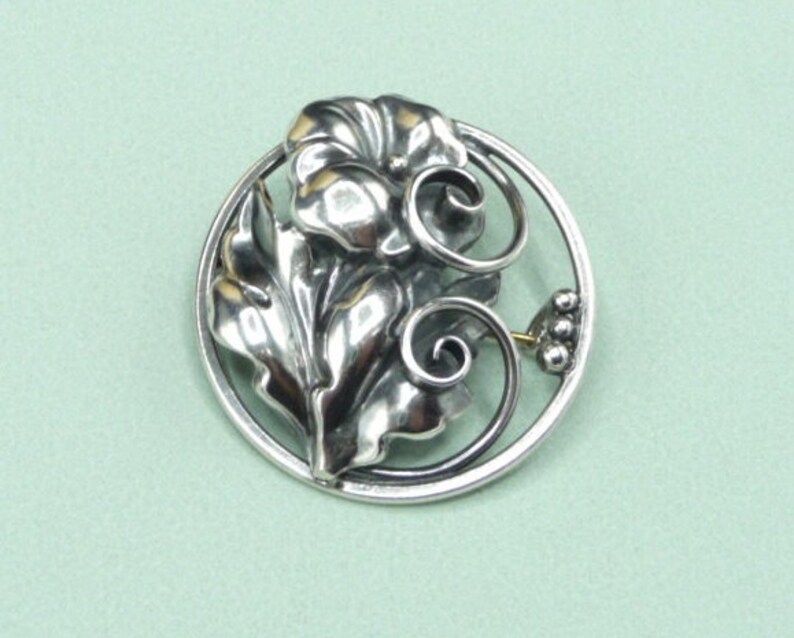 May include: A silver-toned floral brooch with a detailed design. The brooch features a central flower with layered petals, surrounded by leaves and swirling accents within a circular frame. A small cluster of beads and a pin clasp are visible.