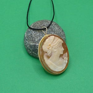 Puede incluir: Un colgante de camafeo ovalado con el perfil tallado de una persona con peinado recogido, engastado en un marco de metal dorado. El colgante cuelga de un cordón negro fino y está colocado frente a un objeto de piedra gris, redondeado y texturizado.