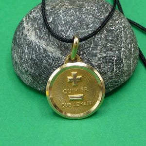 May include: A round gold-tone pendant on a black cord necklace. The pendant features a raised cross symbol, a minus sign, and the French text QU'HIER QUE DEMAIN engraved on the textured gold surface. The design has a polished gold border.