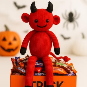 Puede incluir: Una muñeca de diablo roja tejida con cuernos, orejas y pies negros está sentada encima de una caja naranja llena de caramelos. La caja dice "TRICK OR TREAT" en letras negras. Una calabaza tallada y decoraciones de Halloween están en el fondo.
