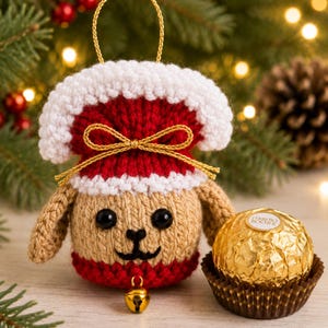 Peut inclure: Une décoration en tricot représentant un chien beige et rouge avec une bordure blanche, des yeux en boutons noirs, un nez brodé et une clochette dorée à la base. Elle possède une boucle dorée et un nœud doré. Un chocolat Ferrero Rocher est posé à côté.