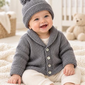 May include: A grey knit cardigan with a shawl collar and four light-coloured buttons, paired with a matching grey knit beanie featuring a large pom-pom on top. The outfit is worn over a plain white shirt and cream-coloured knit trousers.