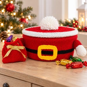 Könnte beinhalten: Ein roter Strickbehälter mit einem weißen Bommel-Deckel, verziert mit einem schwarzen Gürtel und einer gelben Schnalle. Dazu gehört ein kleiner, passender roter Strickbeutel mit goldenem Band sowie mehrere in rotes, grünes und goldenes Papier eingewickelte Bonbons.