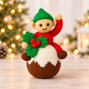 May include: A knitted doll featuring a tan face with black eyes and a small smile, wearing a green hat and red clothing. The doll sits atop a brown and white pudding-shaped base, holding a green holly leaf decoration with three red berries.