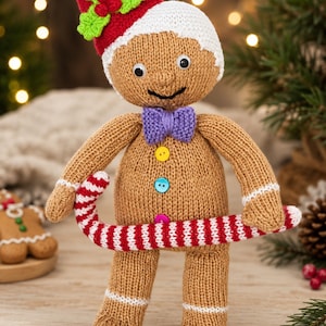 Puede incluir: Una muñeca de punto con forma de hombre de jengibre color canela, con un gorro rojo con hojas de acebo verdes y bayas rojas, pajarita morada, tres botones de colores y ribetes blancos en brazos y piernas, sosteniendo un bastón de caramelo.