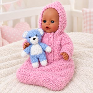 May include: A doll with blue eyes and a light skin tone wearing a pink hooded, long-sleeved knitted outfit with three buttons down the front. The doll is holding a small, light blue and white knitted teddy bear with a black nose and eyes.