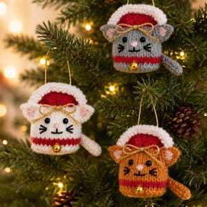 Op de afbeelding: Drie gebreide kattenornamenten in grijs, wit en oranje, elk met een rood-witte muts met een gouden strik en een klein gouden belletje aan de onderkant. Elke kat heeft geborduurde gelaatstrekken, puntige oren en een staartje aan de zijkant.