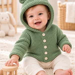 May include: A sage green knit hooded cardigan featuring two small rounded ears on top of the hood and five light-colored wooden buttons down the front. The long-sleeved garment is crafted with a textured garter stitch pattern and includes a hood.