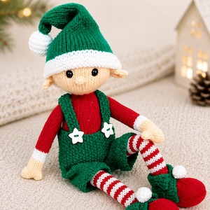 Pode incluir: Uma boneca elfo de malha com um gorro verde com um pompom branco, uma camisa vermelha de manga comprida e jardineiras verdes com dois botões brancos em forma de estrela. A boneca tem pernas às riscas vermelhas e brancas e sapatos verdes.