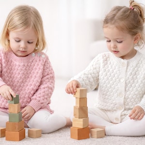 May include: Two young children are playing with wooden blocks. One child wears a pink sweater, the other a white cardigan. The blocks are various shades of brown and green, stacked into towers. The children are sitting on a light-colored surface.