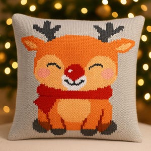 May include: A square, knitted cushion featuring a smiling cartoon reindeer. The reindeer has orange fur, a red nose, and a red scarf. The background of the cushion is light grey, and the cushion is decorated with a Christmas theme.