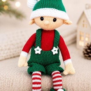 May include: A knitted elf doll featuring a green hat with a white pom-pom, a red long-sleeved shirt, and green overalls with two white star-shaped buttons. The doll has striped red and white legs and green shoes with white pom-poms at the toes.