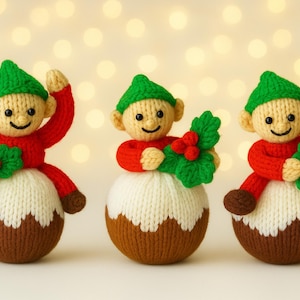以下が含まれることがあります： 3つの編み物で作られたクリスマスのエルフのオーナメント。それぞれのエルフは緑の帽子と赤いシャツを着て、茶色、白、茶色のプディングの上に座っています。エルフは赤い実のついた緑のヒイラギを持っています。背景は柔らかくぼやけたボケです。
