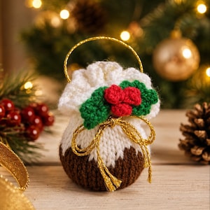 May include: A small, round, crocheted pouch designed to resemble a Christmas pudding. The base is dark brown, topped with white yarn, and decorated with three red berries and two green leaves. A gold metallic cord forms a drawstring and handle.