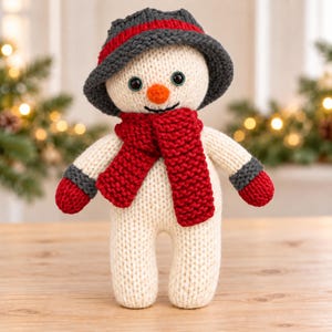 May include: A hand-knitted snowman doll made of cream-coloured yarn, featuring a dark grey hat with a red stripe, a bright red scarf, and matching red mittens. The snowman has black button eyes and a small orange nose, standing upright.