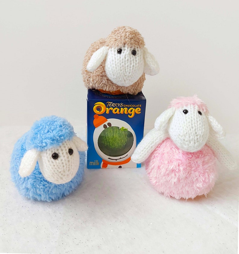 K&ouml;nnte beinhalten: Drei gestrickte Schafspielzeuge, eines blau, eines rosa und eines braun, die auf einer wei&szlig;en Oberfl&auml;che liegen. Das braune Schaf sitzt auf einer Schachtel Terry's Chocolate Orange.