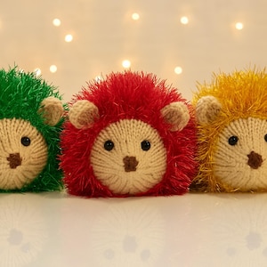 Può includere: Tre peluche di riccio fatti a mano in fila. I ricci hanno facce lavorate a maglia con occhi neri e nasi marroni. Uno è verde, uno rosso e uno giallo, con pelliccia soffice abbinata. Lo sfondo è sfocato con luci soffuse e calde.