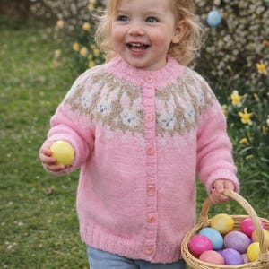 Puede incluir: Un cárdigan rosa con un diseño de conejitos en el canesú, usado por un niño. El niño sostiene un huevo amarillo y una cesta llena de coloridos huevos de Pascua. El cárdigan tiene botones.