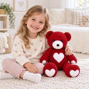 Op de afbeelding: Een pluche teddybeer gemaakt van donkerrood garen, met een witte snuit, zwarte knoopogen en een zwarte neus. De beer heeft een groot wit hart op de borst en kleinere witte hartjes op elk van de twee voeten.