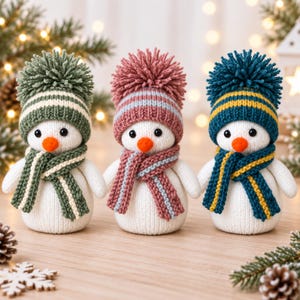 May include: Three white knit snowman figurines, each wearing a matching pom-pom hat and scarf. The first snowman wears green and white, the second wears dusty pink and grey, and the third wears dark blue and yellow. Each has a small orange nose.