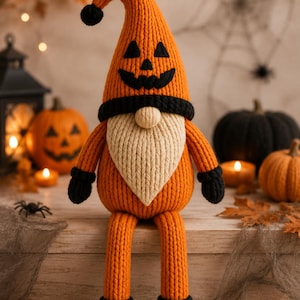 May include: A knitted gnome figurine featuring a tall orange hat with a black jack-o'-lantern face, a beige beard, long orange legs, and black mittens and boots. The gnome has a round beige nose and a black band separating the hat from the face.