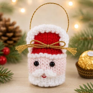 May include: A small, hand-knitted Santa Claus ornament featuring a red hat with white trim, a pale pink face, black bead eyes, a white moustache, and a small red mouth. It includes a gold metallic hanging loop and a gold ribbon bow.